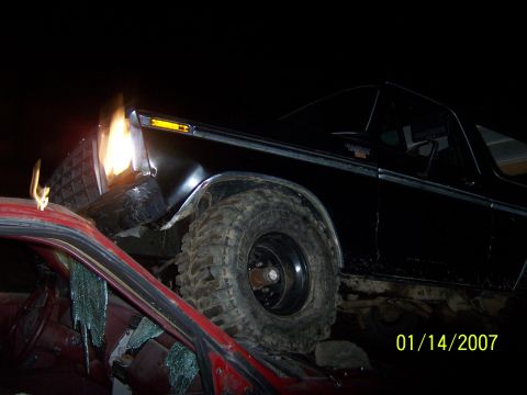 1989 Ford Bronco - A black Ford Bronco is heavily damaged and appears to be on fire at the front bumper. The vehicle is resting on top of a red car, with brok…