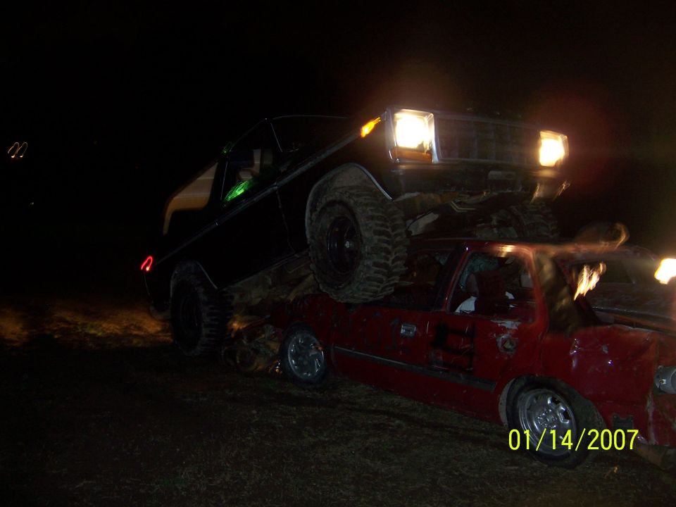 Black SUV on top of a red sedan at night, SUV headlights on.