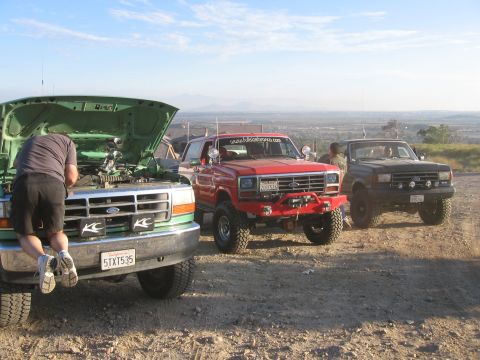 1996 Ford Bronco - A person works on the engine of a green Ford Bronco with the hood open, parked on a dirt road with two other Broncos behind it.