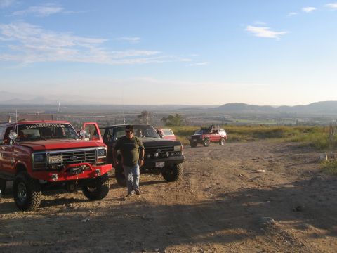 1996 Ford Bronco - A man stands next to a red Ford Bronco with a custom front bumper and winch. The vehicle is parked on a dirt lot with other off-road vehicl…