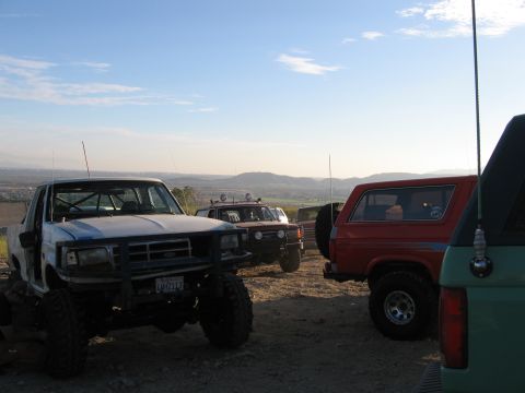 1996 Ford Bronco - Several off-road vehicles are parked on a dirt lot with a scenic view of hills and a town in the distance under a partly cloudy sky.