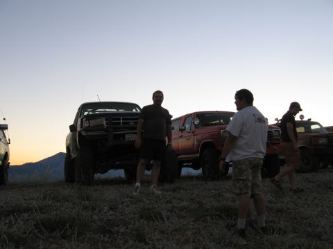 1996 Ford Bronco - A group of people stand around several lifted off-road vehicles parked on a grassy hill at dusk. The vehicles include a dark-colored pickup…