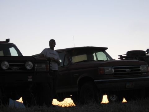 1996 Ford Bronco - A man leans against the hood of a dark-colored Ford Bronco parked in a grassy field at dusk. The vehicle's headlights and grille are visibl…