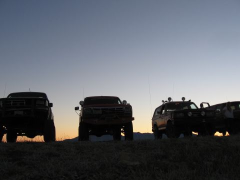 1996 Ford Bronco - Several off-road modified trucks and SUVs are parked on a grassy hill against a gradient sunset sky. The vehicles are silhouetted, with the…
