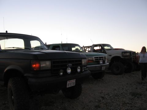 1996 Ford Bronco - A line of modified pickup trucks and SUVs are parked on a dirt lot at dusk. The foreground vehicle is a dark-colored pickup truck with larg…