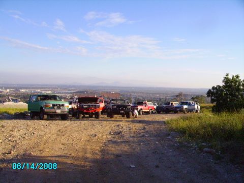 1990 Ford Bronco - A group of off-road vehicles are gathered on a dusty hilltop, overlooking a urban landscape under a bright blue sky.