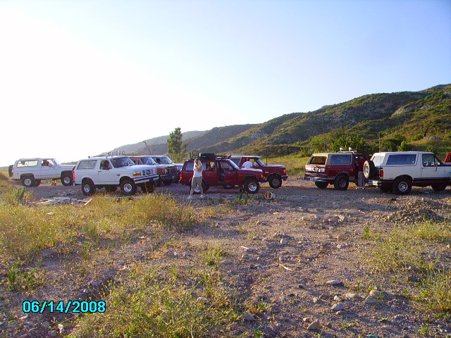 Several Ford Broncos and other SUVs parked on a dirt lot with hills in the background. 1990 Ford Bronco is among them