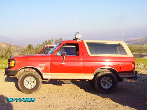1990 Ford Bronco - Side view of a red 1990 Ford Bronco with a tan removable top, parked on a dirt road.