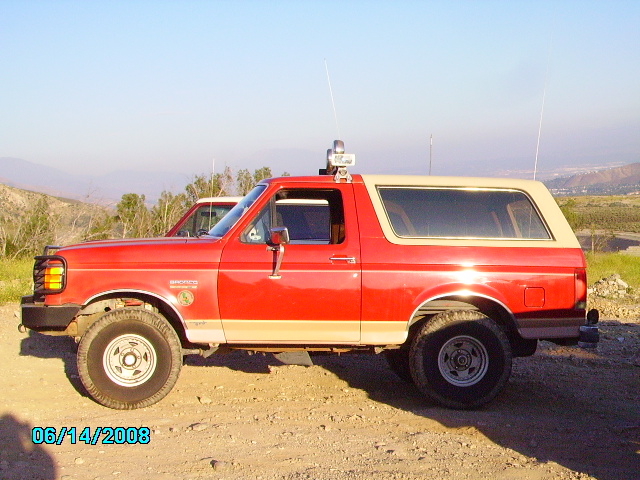 1990 Ford Bronco side profile, red with tan top, parked outdoors with another vehicle behind it
