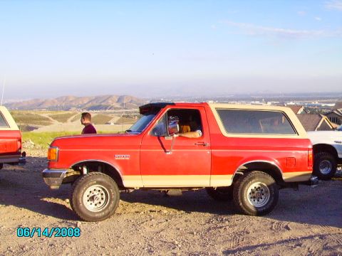 1990 Ford Bronco - A red Ford Bronco with a tan top is parked on a dirt lot with a man standing nearby and another vehicle behind it.