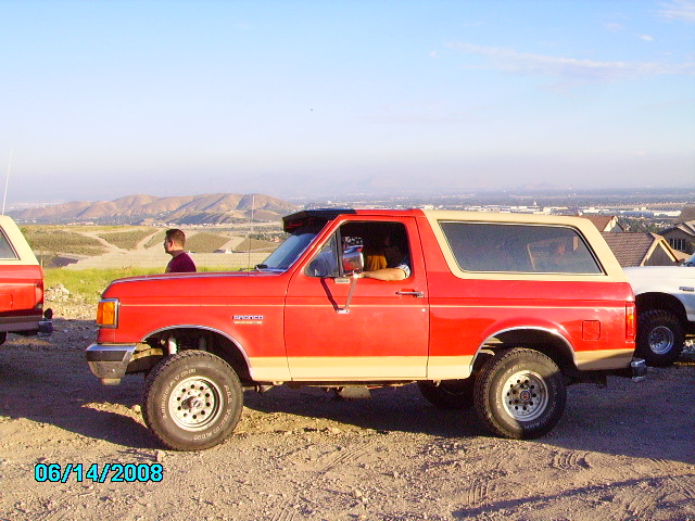 Red Ford Bronco with tan removable top, side view, lifted suspension, off-road tires, parked on dirt with distant hills
