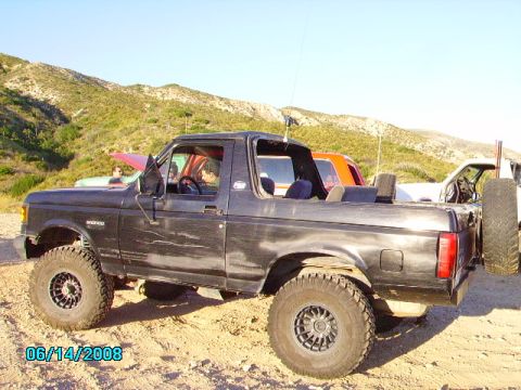 1990 Ford Bronco - This black Ford Bronco is parked outdoors with other vehicles and a hilly landscape in the background.