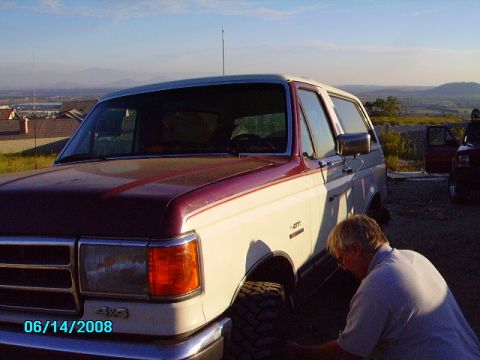 1990 Ford Bronco - A man inspects the front tire of a two-tone Ford Bronco parked outdoors with a scenic view in the background.