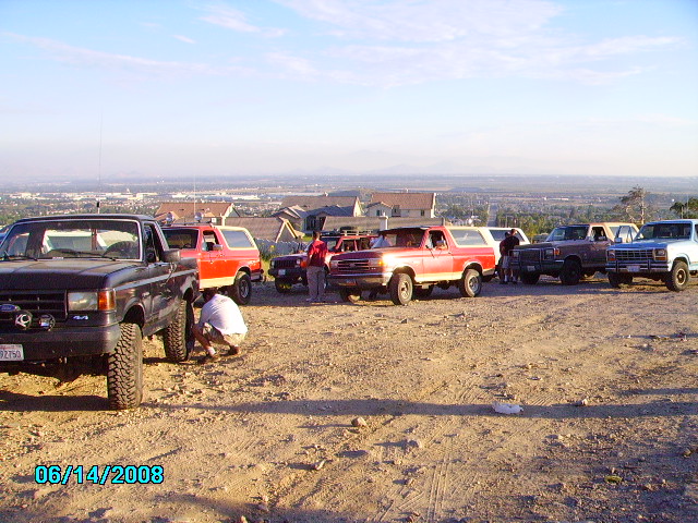 Several Ford Broncos and trucks parked on a dirt lot overlooking a town. One person is working on a tire