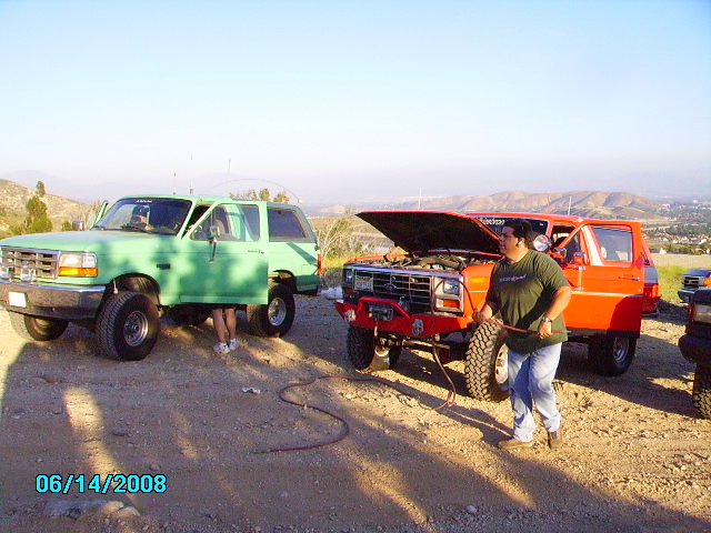 Green Ford Bronco parked next to an orange Ford Bronco with its hood up. A person is standing by the orange Bronco