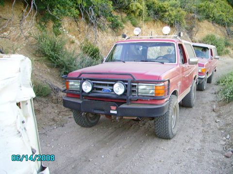 1990 Ford Bronco - Two red SUVs, one a 1990 Ford Bronco, drive on a dusty trail with scrub brush on the side.