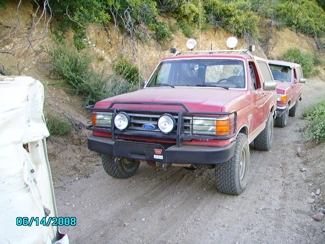 1990 Ford Bronco with a brush guard and auxiliary lights, followed by another vehicle on a dirt road