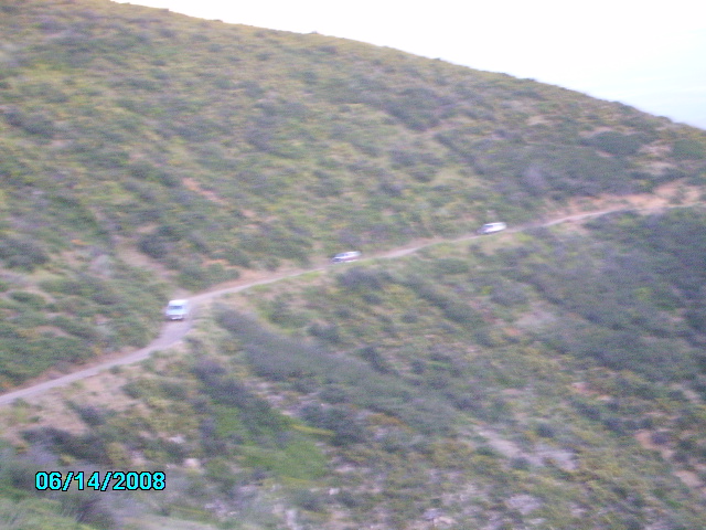 Four vehicles drive along a narrow dirt road winding up a scrub-covered hillside. Date stamp 06/14/2008