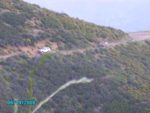 1990 Ford Bronco - Two SUVs navigate a narrow dirt road winding up a vegetated hillside under a hazy sky.