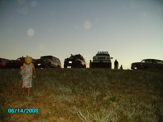 Several SUVs and trucks parked on a grassy hill at dusk. A child in a dress and hat stands in the foreground