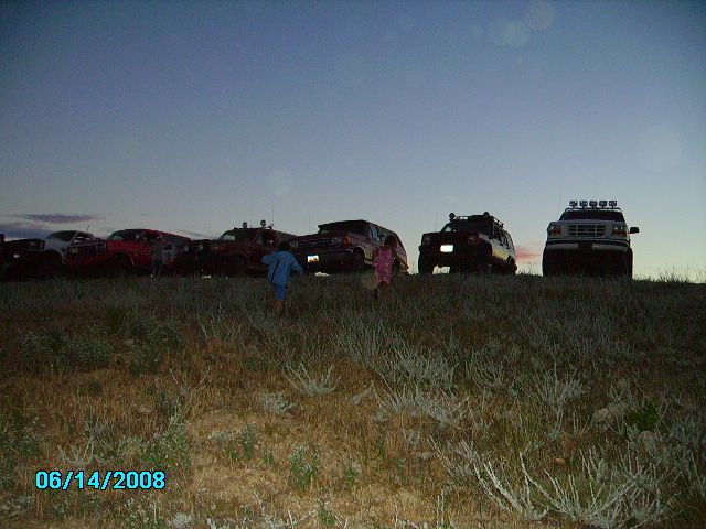 Several off-road vehicles parked on a hill at dusk, with two children walking in the foreground