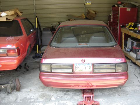 1988 Ford Bronco - Two Ford Mustangs are parked in a garage, one is raised on a jack with its rear end visible.