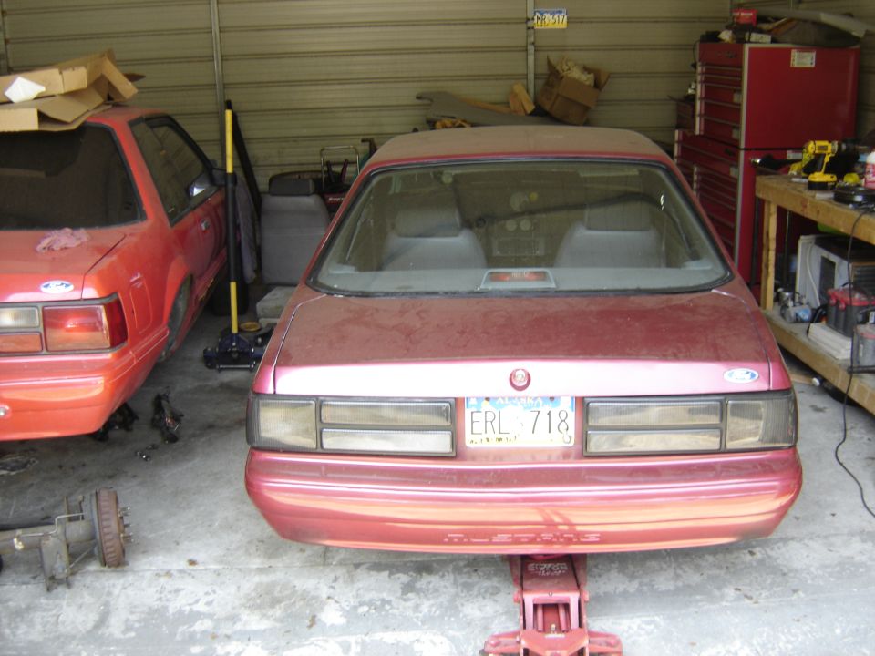 Rear view of a pinkish-red Ford Mustang on a jack, another red Mustang in the background