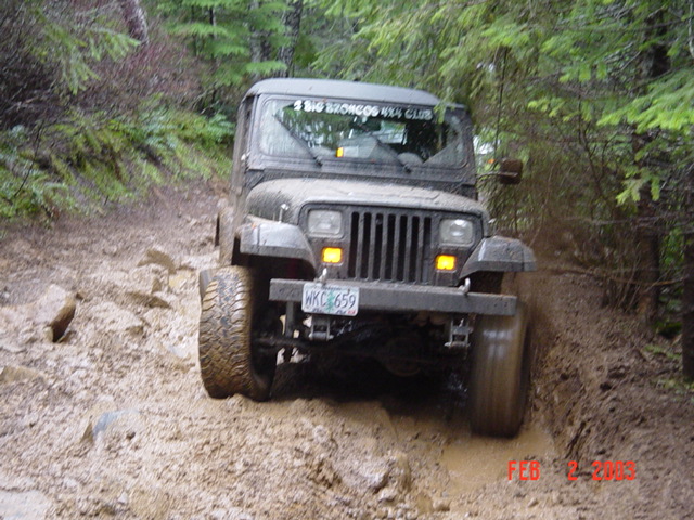 1995 Jeep Wrangler driving on a muddy, rocky trail in a wooded area. License plate reads WKC 659