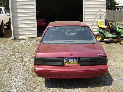 1988 Ford Bronco - Rear view of a red coupe, possibly a Ford Mustang, parked outside a garage with other vehicles present.