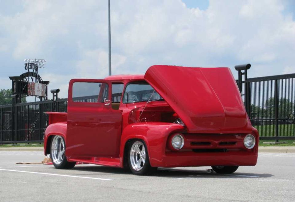 Red customized 1956 Ford F-100 pickup with raised hood and chrome wheels in a parking lot