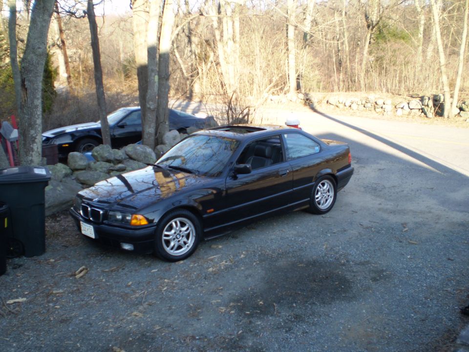 Black BMW coupe with sunroof parked on gravel, front 3/4 view. Another car is visible behind it