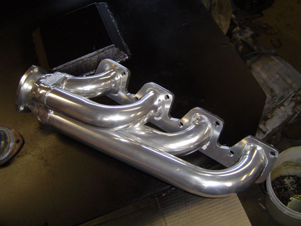 Polished metal exhaust manifold with four pipes merging into two, resting on a black surface