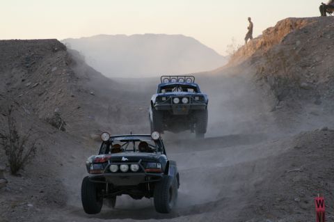 1996 Ford Bronco - Off-road trucks compete in a desert race, kicking up dust as they climb a hill.