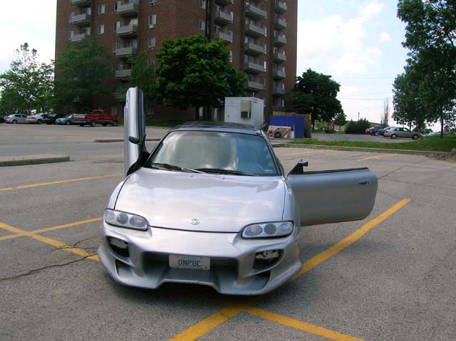 Silver Mazda MX-6 coupe with doors open, aftermarket bumper, plate ONPOC, parked before apartment tower