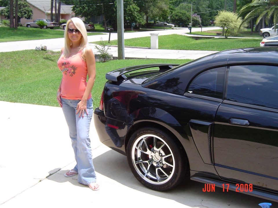 Black Mustang coupe rear quarter with polished deep-dish wheels, woman standing beside it