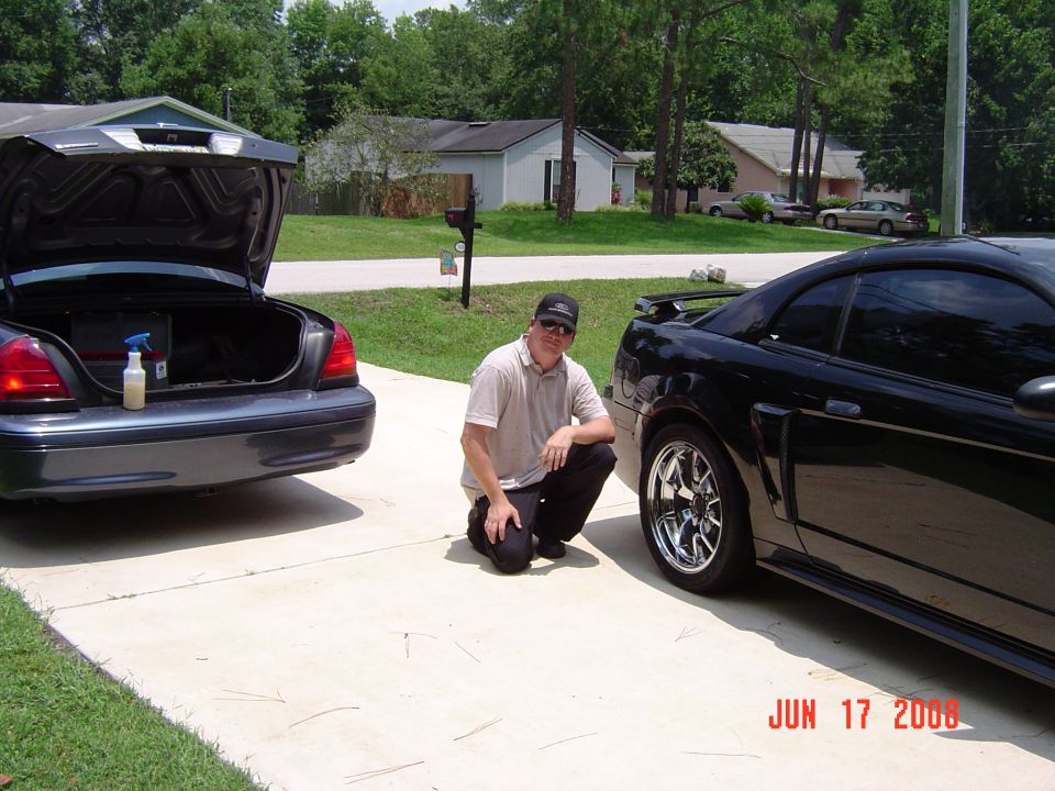 Man kneeling by black Mustang coupe with chrome wheels next to a dark sedan on a driveway