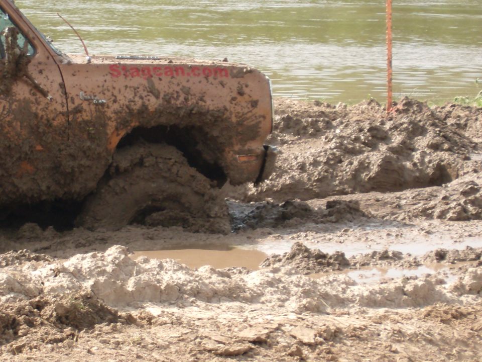 Orange truck stuck in mud, front wheel deep in mire, with "Stagecan.com" visible on the door