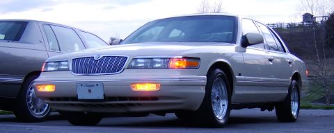 1996 Mercury Grand Marquis - A tan 1996 Mercury Grand Marquis is parked next to another vehicle, showing its front grille and headlights.