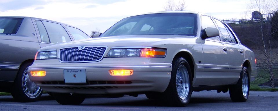 Champagne full-size sedan, front 3/4 view, lights on, chrome grille and wheels, Maryland plate