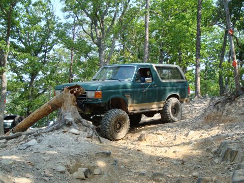 1990 Ford Bronco - A green Ford Bronco navigates a challenging off-road course, climbing over a large tree trunk.