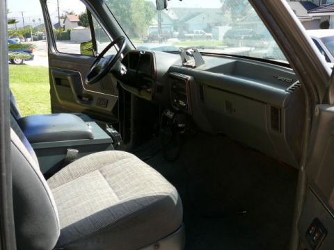 1988 Ford Bronco - Interior of an older Ford Bronco shot from the passenger side through the open door, showing the gray dash with 'Bronco' badging on the glo…