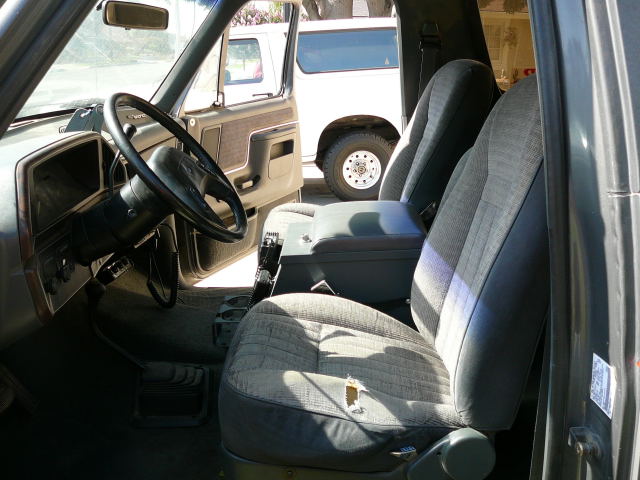 1988 Ford Bronco interior showing ripped driver's seat, dashboard, and steering wheel. Another Bronco is visible outside