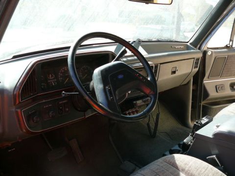 1988 Ford Bronco - Driver-side interior view of a Ford Bronco showing the red/maroon dashboard, two-spoke black steering wheel with blue Ford oval, round anal…