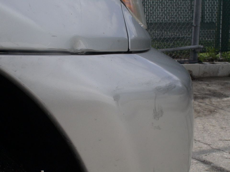 Silver car bumper and fender with scuffs and a dent near the headlight
