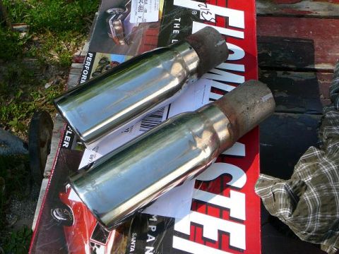 1997 Mercury Grand Marquis - Two chrome angle-cut exhaust tips with rusty weld-on inlet necks lying on a red and black Flowmaster muffler box outdoors. A barcode label…