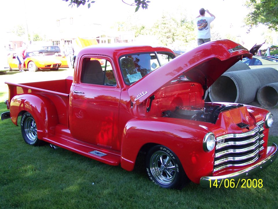 Red 1950 Chevrolet 3100 pickup truck with hood open, parked on grass. Another yellow car is visible in the background