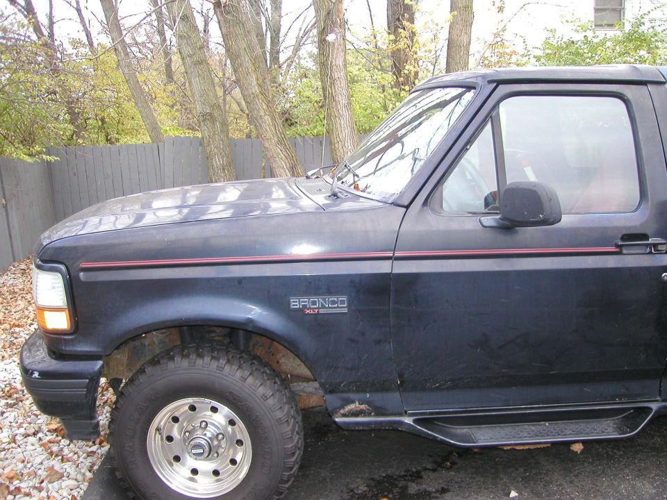 Black Ford Bronco XLT with red pinstripe and chrome wheels, side view by wooden fence and trees