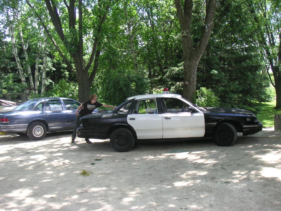 Side view of black-and-white Crown Victoria police sedan being worked on in shaded gravel drive