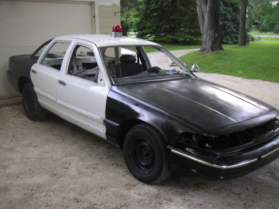 Stripped black-and-white Crown Victoria police sedan with red roof beacon on gravel drive