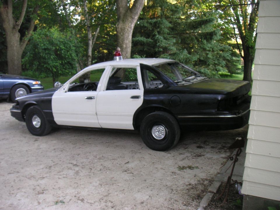 Black-and-white Crown Victoria police car stripped of doors with red roof beacon on gravel drive
